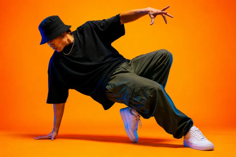 Danseur hip hop en tenue streetwear oversize réalisant une figure de breakdance sur fond orange vif avec éclairage contrasté.